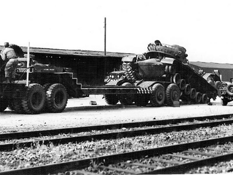 Ein amerikanischer Panzer fährt auf die Ladefläche eines Trucks. Im Hintergrund ein Jeep und Lagerhallen