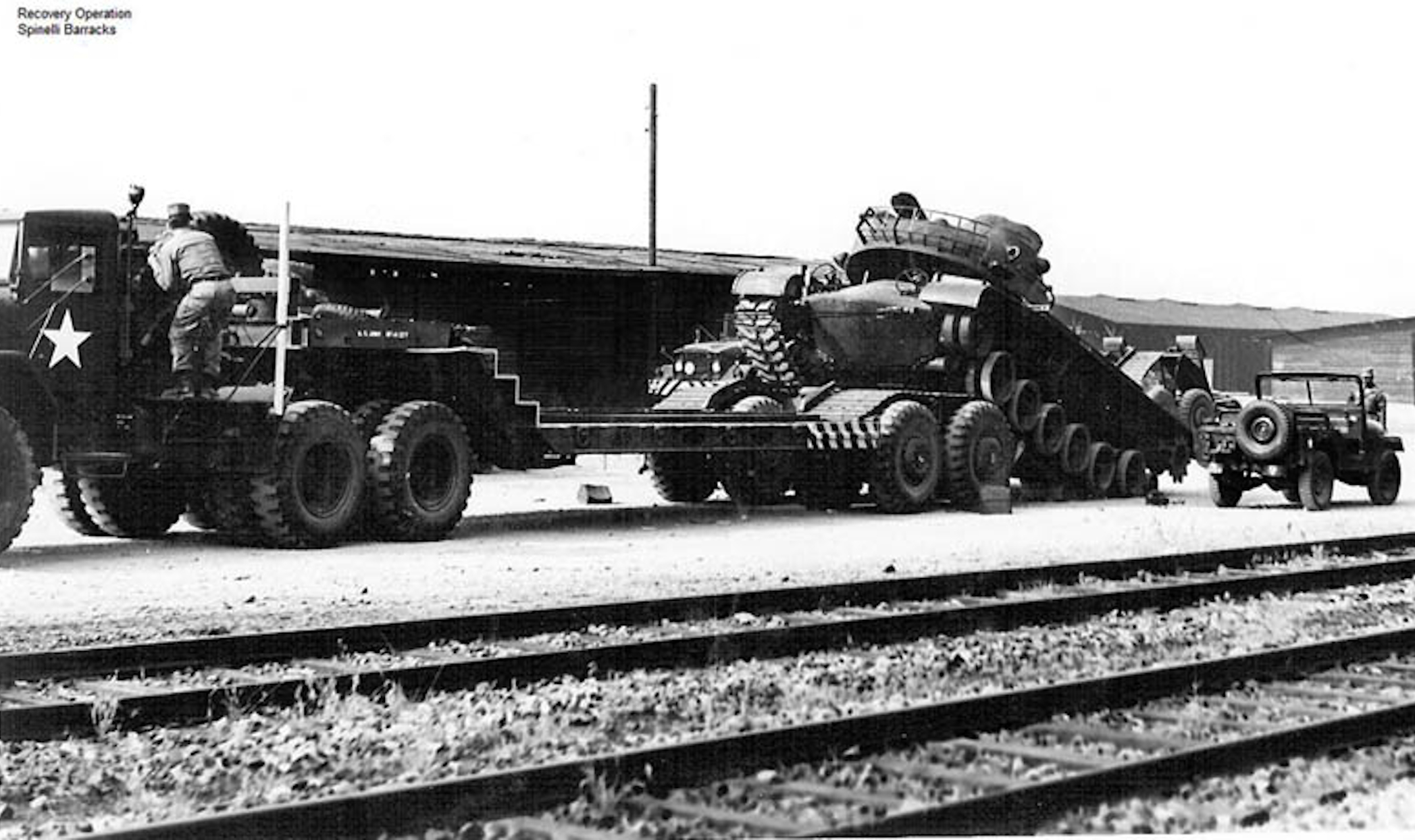 Ein amerikanischer Panzer fährt auf die Ladefläche eines Trucks. Im Hintergrund ein Jeep und Lagerhallen