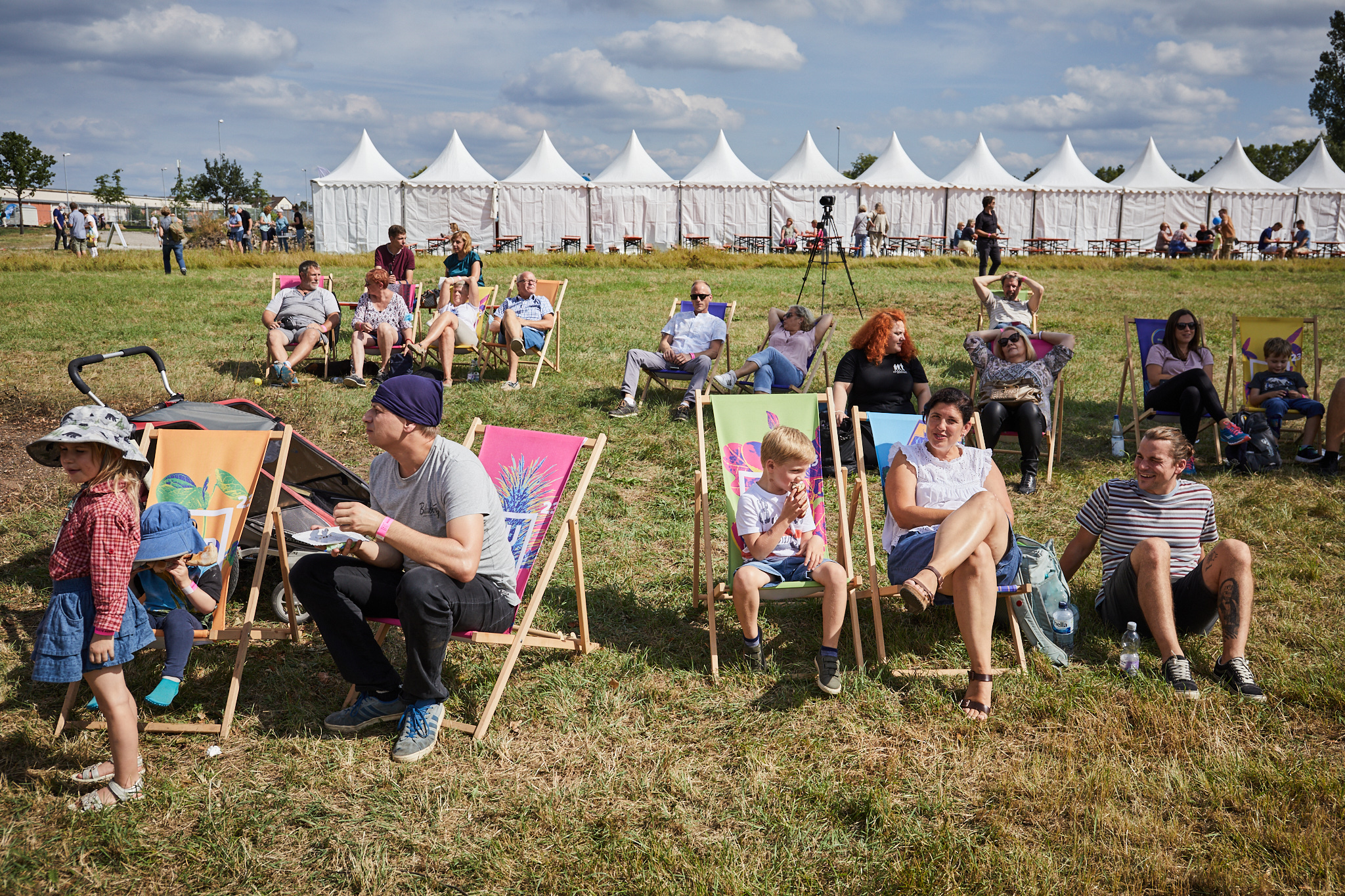 Menschen sitzen im Sommer auf Bierbänken und Liegestühlen. Im Hintergrund Zelte