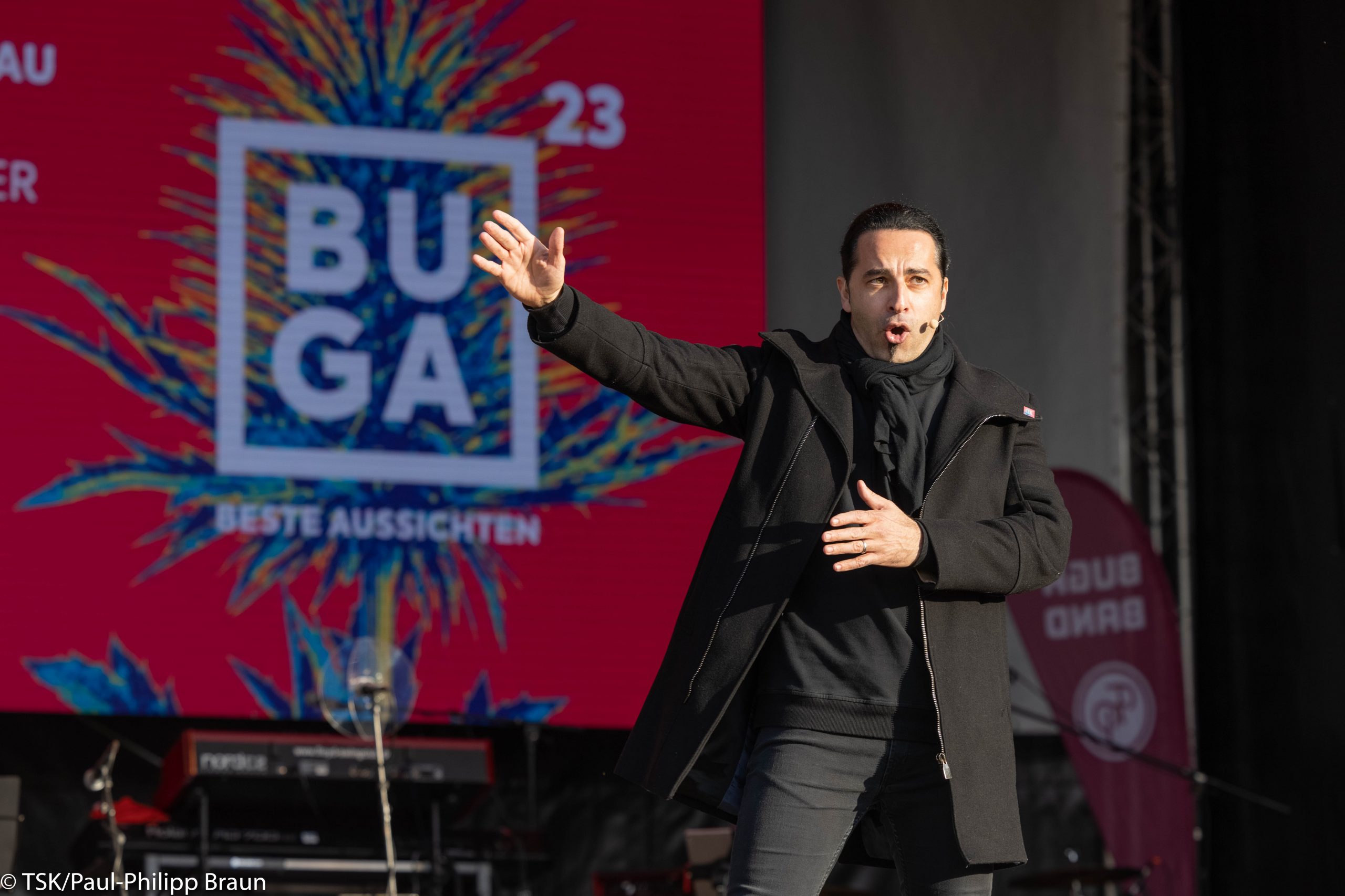 Bülent Ceylan in schwarzem Mantel auf der Bühne. Hinter ihm das Buga 23 Logo