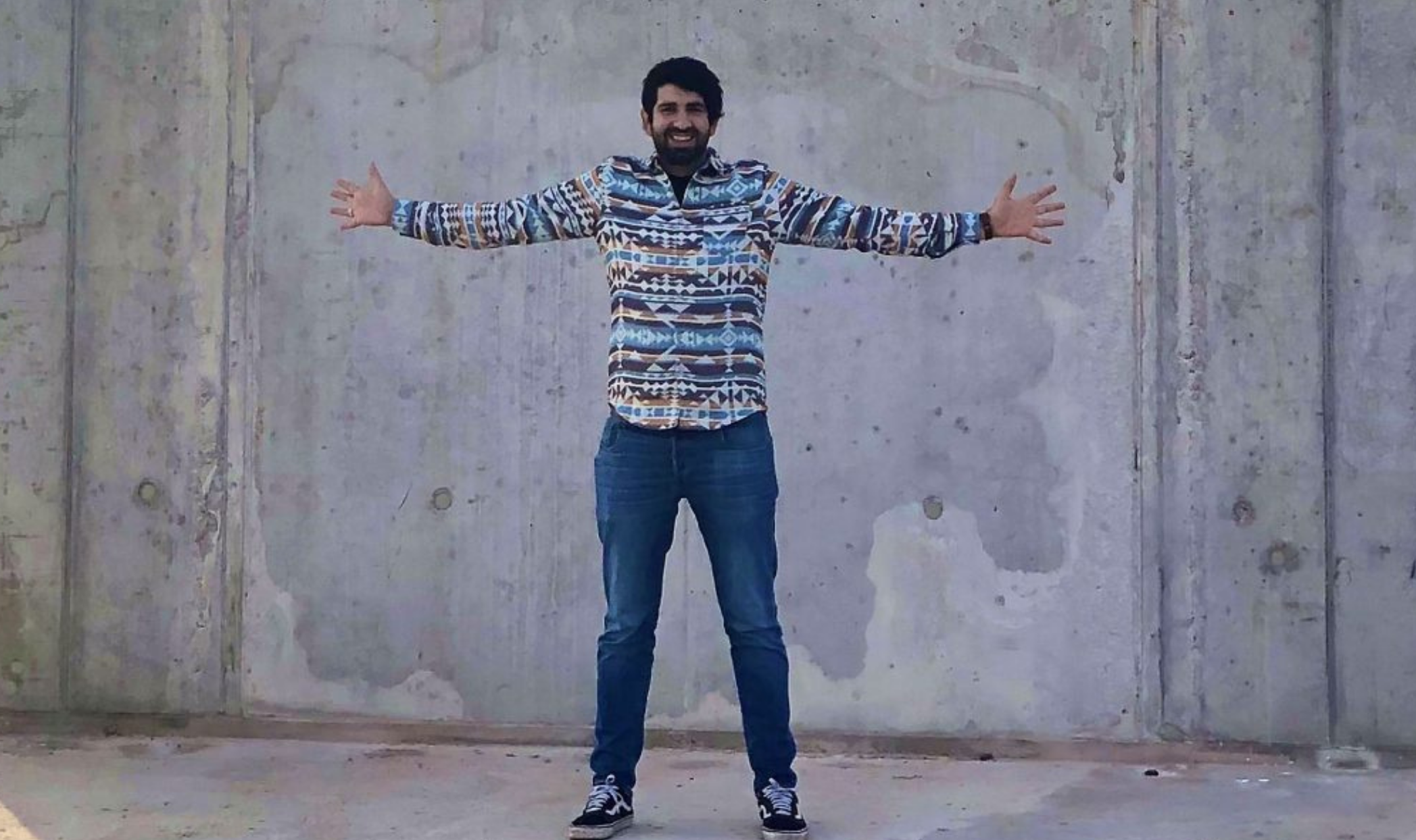 Farsad Tawakol steht mit stylischem sweater und ausgestreckten Armen vor einer Betonwand