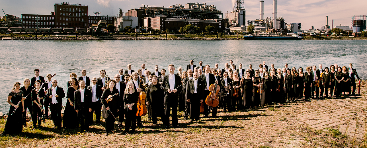 Ein Orchester steht an einem Flussufer. Am gegenüberliegenden Ufer befindet sich eine Fabrik