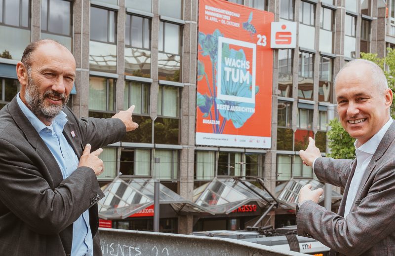 Der Geschäftsführer der BUGA 23 gGmbH Michael Schnellbach und der Vorstandsvorsitzende der Sparkasse Rhein Neckar Nord zeigen auf ein Rotes Buga-Banner, das an einer Hauswand hängt