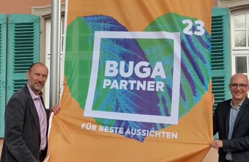 Buga-Geschäftsführer Michael Schnellbach und Schwetzingens Oberbürgermeister René Pöltl Halten die BUGA-Partnerflagge in der Hand