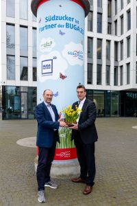 BUGA 23 Geschäftsführer Michael Schnellbach und Dr. Niel Pörksen stehen vor einer Säule und halten einen gelben Blumenstrauß