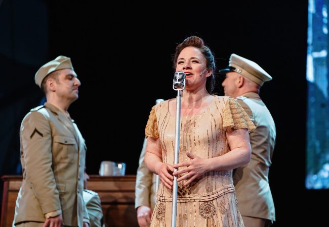 Die Hauptdarstellerin des Musicals Jeannette Friedrich (als Joy Fleming) steht am Mikrofon. Um sie herum stehen amerikanische Soldaten