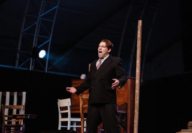 Ein Mann in schwarzem Nadelstreifenanzug singt, hinter ihm stehen Holzmöbel