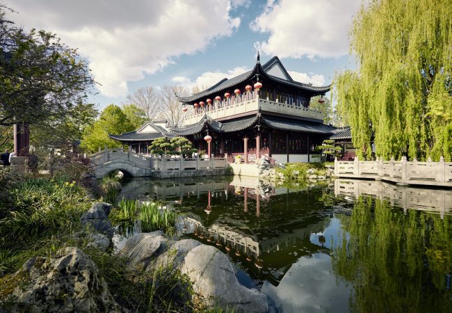 Ein Chinesisches Teehaus umgeben von Bäumen. Im Vordergrund ein Teich