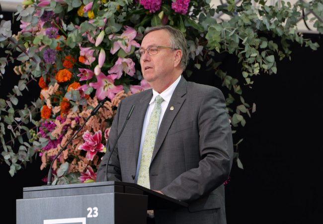 Hajo Hinrichs steht am Rednerpult der Hauptbühne. Im Hintergrund Blumen