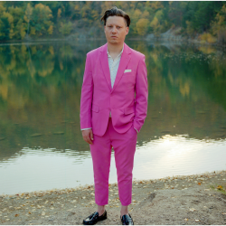 Konstantin Gropper (in pinkem Anzug) steht vor einem See und blickt in die Kamera